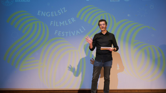 In front of a blue stage background, a man dressed in a black shirt and dark jeans is speaking while holding a piece of paper and gesturing with his right hand. On the blue backdrop, the number 2026 is formed by yellow and green stripes, positioned next to the text Engelsiz Filmler Festivali. At the very top of the blue background, various institutional and organizational logos are displayed.