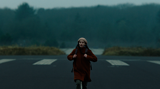 In the film photograph, a young woman is walking towards the camera on a wide asphalt ground, holding the straps of her bag on her shoulder. The young woman is wearing a burgundy coat and gray pants. She has a woolen beanie on her head. The forested terrain beyond the asphalt ground fills the entire background of the photograph. A closed sky occupies a small portion at the top of the frame.
