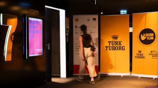 A young woman with long hair walks towards the door of movie theater number 7. to the left of the door hangs the poster of the Accessible Film Festival. to the right of the door are roll-ups with the names of türk tuborg, sesim elim and festival sponsors.