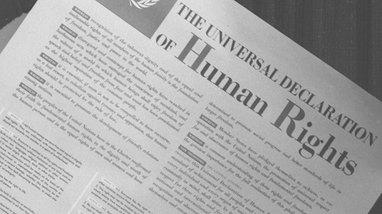 The newspaper in the black and white photograph has English writing on it. The newspaper reads THE UNIVERSAL DECLARATION OF HUMAN RIGHTS in large font. The lower text cannot be read and the frame does not show the entire newspaper page.