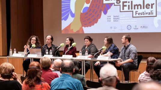 The image depicts a panel discussion with six speakers seated behind a long table in front of a large screen displaying the Accessible Film Festival logo. In the foreground, an audience is seen from behind, with some members wearing headphones for simultaneous translation or audio description. On the left, a female panelist is actively speaking into a microphone using hand gestures, while the other participants listen attentively next to their nameplates and water bottles.
